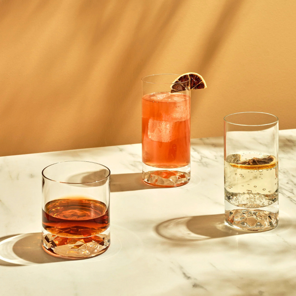 Three drinks served in the Nude Glassware Club Drinkware Set of 4—dark brown, tall pink cocktail with citrus, and clear bubbly with lemon—sit on a marble surface casting elegant shadows on a beige background.