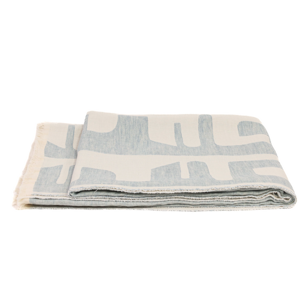 The Uniquity Waters Throw Blue is a folded Belgian linen blanket in light blue and white with an abstract geometric pattern and fringe, shown against a white background.