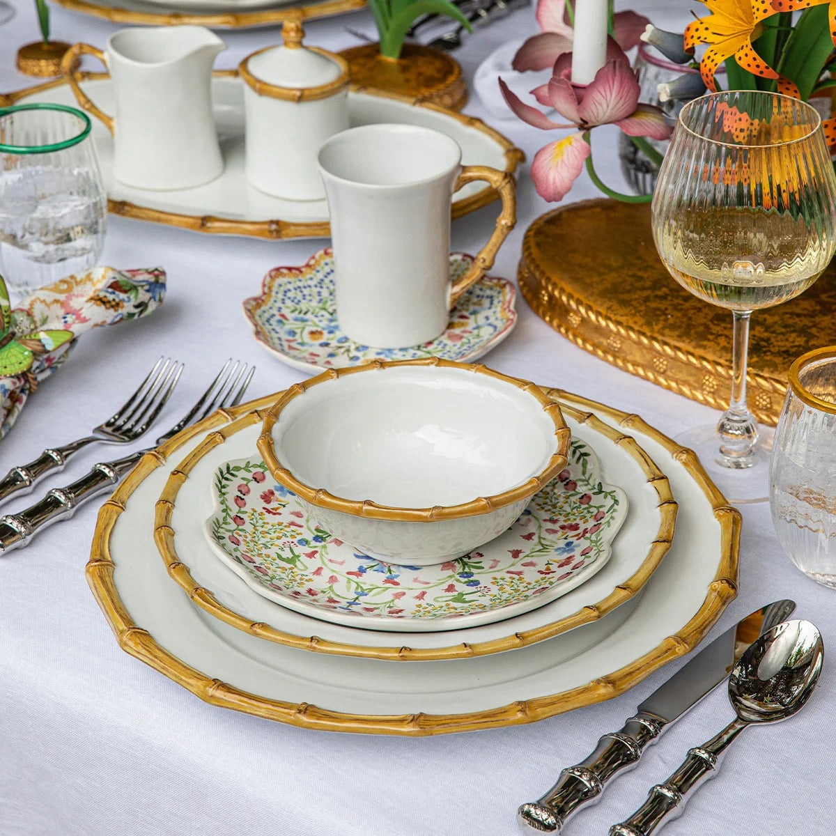 Juliska’s Bamboo Dinnerware creates an elegant table setting with its white and floral-patterned design, complemented by gold-rimmed edges. The arrangement features glassware, silver cutlery, decorative flowers on a white tablecloth, a gold serving platter, and a wine glass to complete the look.