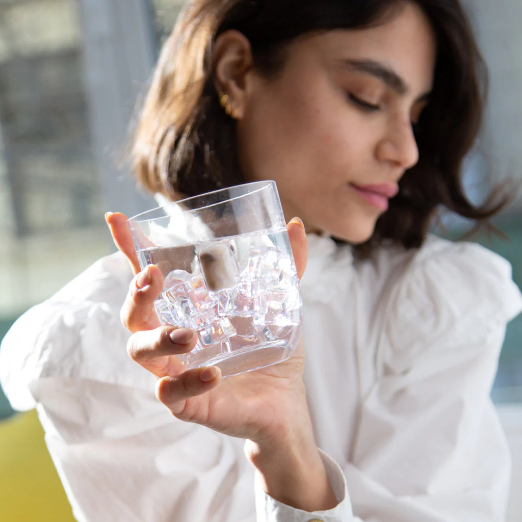 A woman in a white blouse holds up an Elevo DOF Glass from the Smoke Set of 2 by Anna New York, filled with ice and water. The focus is on the elegant glass, while she appears slightly out of focus and turned to the side.