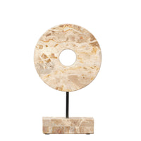 The Jamie Young Marble Cream Circle Disk on Stand features a round, cream marble sculpture with a central hole, set on a black metal stand and rectangular stone base—an elegant, celestial-inspired decor piece.