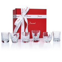 A red Baccarat gift box with a white ribbon sits behind the Take A Shot - Les Mini Set by Baccarat—six crystal whiskey glasses, each featuring a unique cut pattern—arranged on a white background.