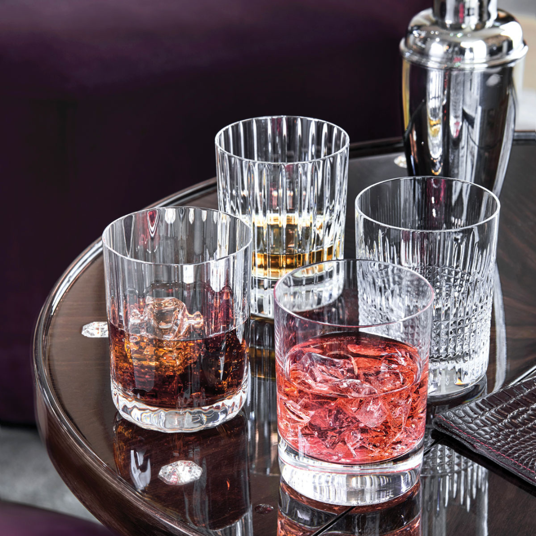 The Baccarat 4 Elements DOF Gift Set features four crystal Double Old Fashioned tumblers, each with a colored drink and ice, arranged on a glossy wooden table beside a metal cocktail shaker and brown mat against a dark purple backdrop.
