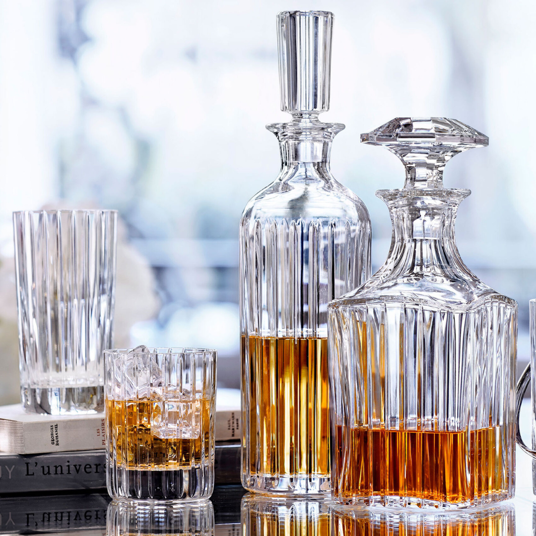 Two Baccarat Harmonie Whiskey Decanter Square bottles, filled with amber liquid, stand beside a whiskey glass with ice, an empty tall glass, and stacked books on a reflective surface against a softly blurred background.