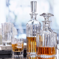 Two Baccarat Harmonie Whiskey Decanter Square bottles, filled with amber liquid, stand beside a whiskey glass with ice, an empty tall glass, and stacked books on a reflective surface against a softly blurred background.