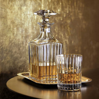 A Baccarat Harmonie Whiskey Decanter Square, partially filled with amber liquor, rests on a small tray beside a matching glass, both placed against a golden textured background.