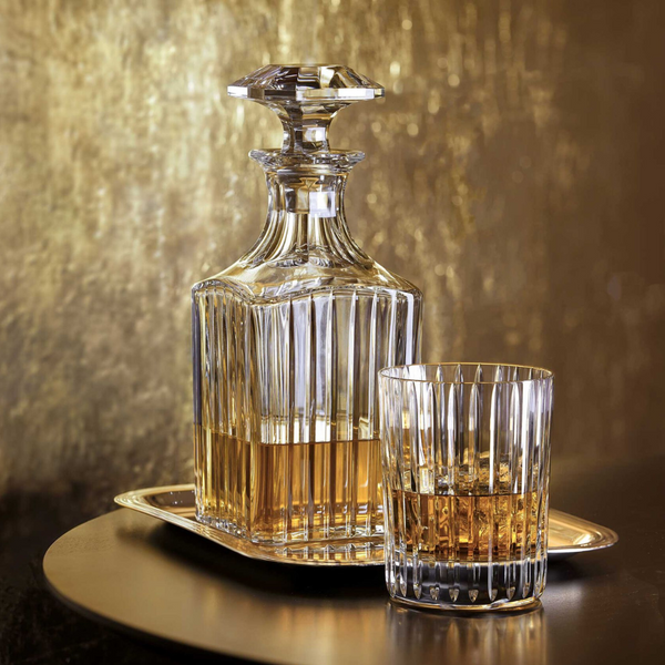A Baccarat Harmonie Whiskey Decanter Square, partially filled with amber liquor, rests on a small tray beside a matching glass, both placed against a golden textured background.