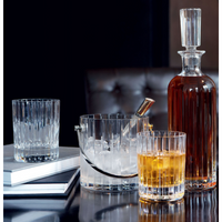 The Baccarat Harmonie Ice Bucket rests beside a glass decanter, two whiskey glasses—one empty, one filled—and stacked books on a glossy table. A leather chair in the background completes this scene of refined barware elegance.