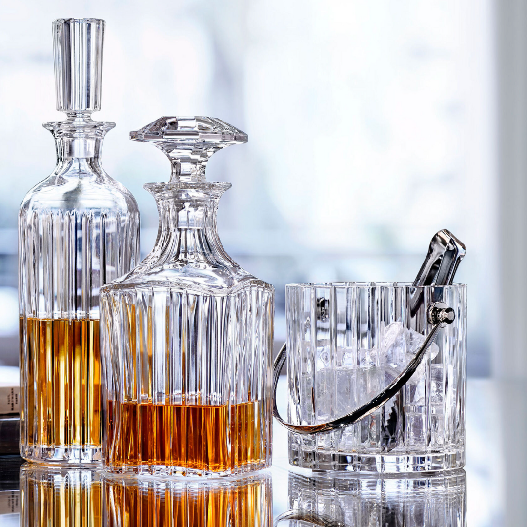 Two crystal decanters filled with amber liquid stand next to the Baccarat Harmonie Ice Bucket with tongs, all arranged on a reflective surface with a softly blurred background suggesting a bright, elegant setting.
