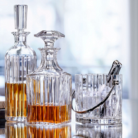 Two crystal decanters filled with amber liquid stand next to the Baccarat Harmonie Ice Bucket with tongs, all arranged on a reflective surface with a softly blurred background suggesting a bright, elegant setting.