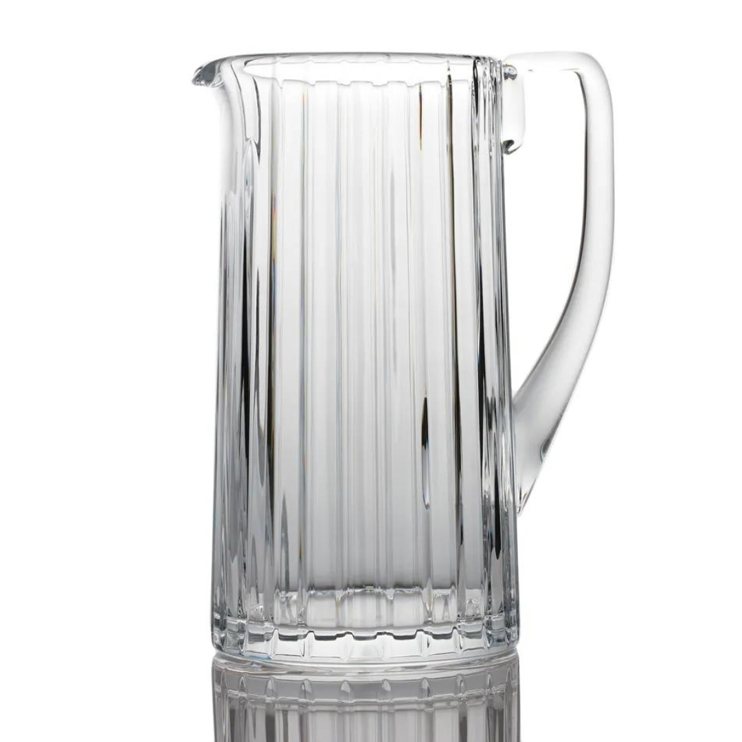 The Baccarat Harmonie Pitcher is a clear crystal piece featuring vertical ridges and a sturdy handle, shown upright on a reflective surface with a white background.