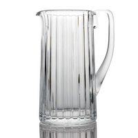 The Baccarat Harmonie Pitcher is a clear crystal piece featuring vertical ridges and a sturdy handle, shown upright on a reflective surface with a white background.