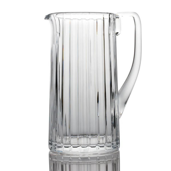 The Baccarat Harmonie Pitcher is a clear crystal piece featuring vertical ridges and a sturdy handle, shown upright on a reflective surface with a white background.