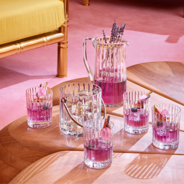 A Baccarat Harmonie Pitcher with pink and purple drinks, plus four glasses garnished with lavender, cinnamon sticks, and ice, rests on a wooden table. A pink floor and yellow chair appear in the background.