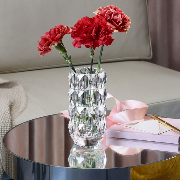 The Louxor Round Vase Small by Baccarat holds three red carnations on a reflective table next to a pink gift box, ribbon, and gold pen, with a beige sofa and blanket in the background.