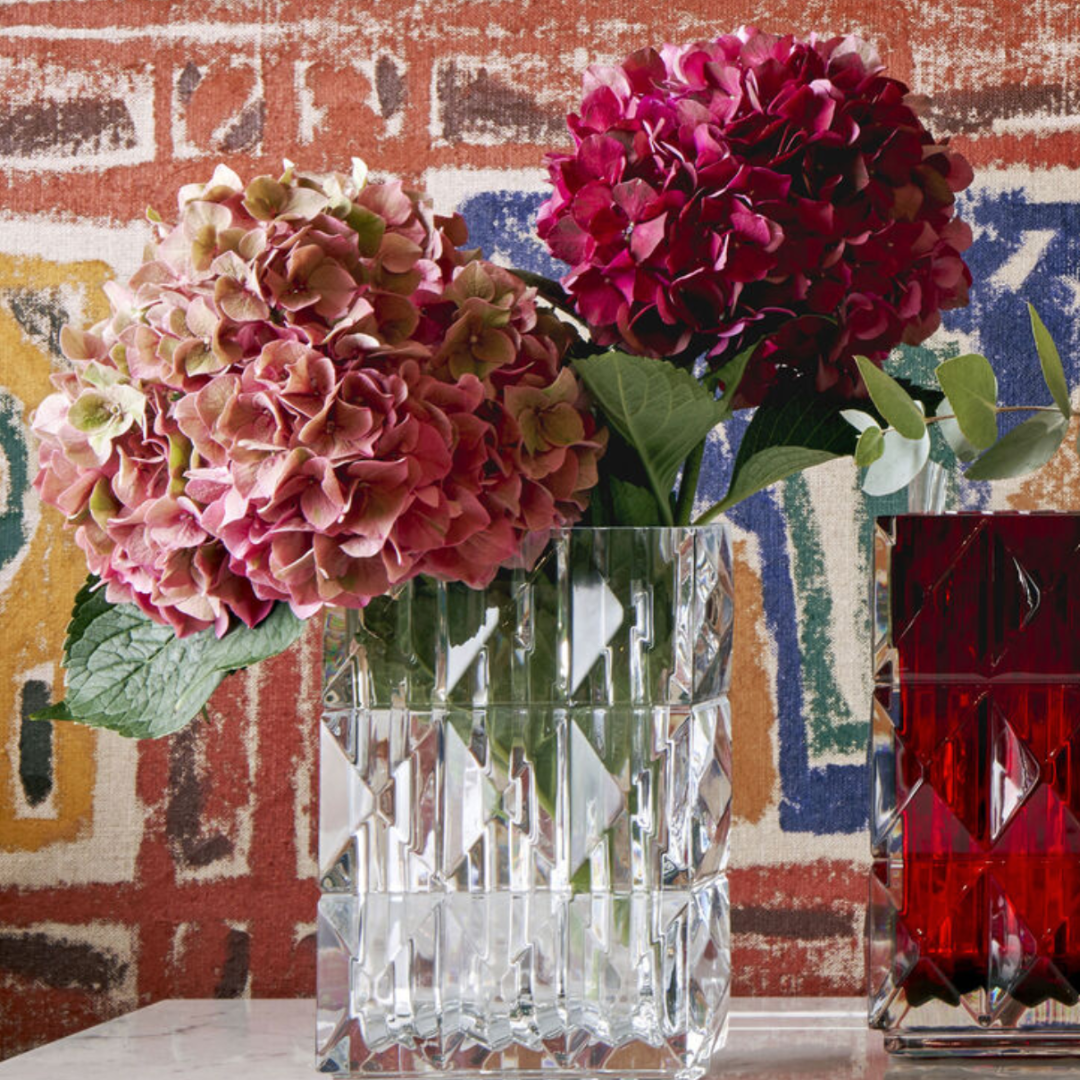 Two pink and red hydrangeas with green leaves are arranged in the Baccarat Louxor Rectangle Vase, set beside a red glass vase against a vibrant geometric wall—an elegant luxury home décor accent.