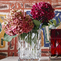 Two pink and red hydrangeas with green leaves are arranged in the Baccarat Louxor Rectangle Vase, set beside a red glass vase against a vibrant geometric wall—an elegant luxury home décor accent.