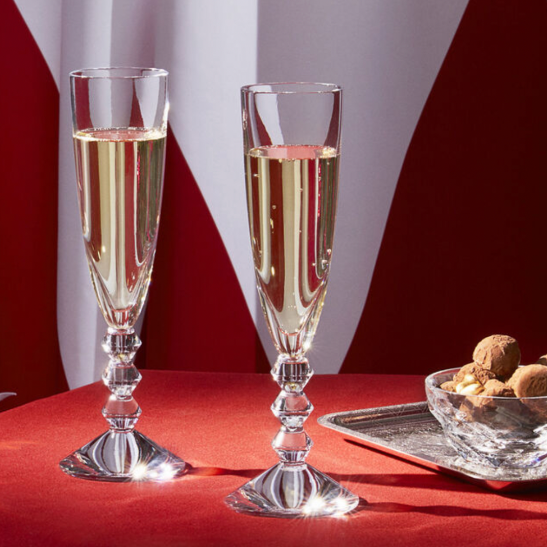 Two Baccarat Vega Champagne flutes, made in France, filled with sparkling wine rest on a red tablecloth next to a glass dish of assorted chocolates and nuts. A red and white patterned fabric appears in the background.