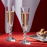 Two Baccarat Vega Champagne flutes, made in France, filled with sparkling wine rest on a red tablecloth next to a glass dish of assorted chocolates and nuts. A red and white patterned fabric appears in the background.