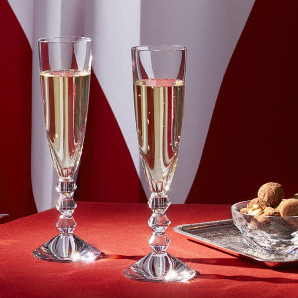 Two Baccarat Vega Champagne flutes, made in France, filled with sparkling wine rest on a red tablecloth next to a glass dish of assorted chocolates and nuts. A red and white patterned fabric appears in the background.