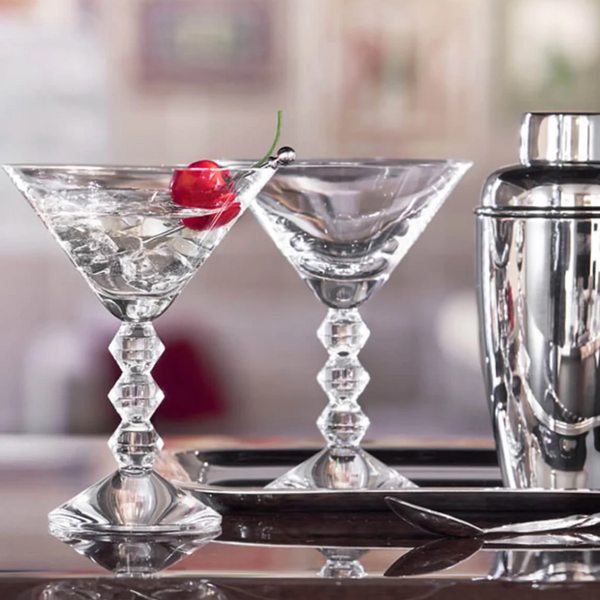 The Baccarat Vega Martini Glass Set of 2, crafted crystal from France, rests on a bar—one glass filled with ice and a cherry garnish, the other empty. A metal shaker, tray, and spoon complete the refined scene.
