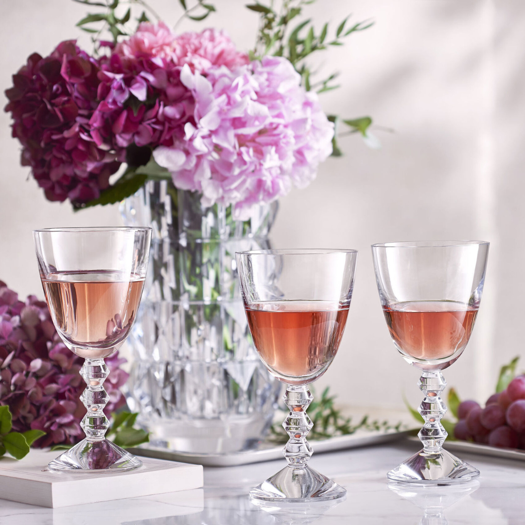 Three Baccarat Vega Red Wine Glasses, crafted in France, are filled with rosé and displayed on a marble surface, accompanied by a crystal vase of pink and purple flowers and grapes in the background.