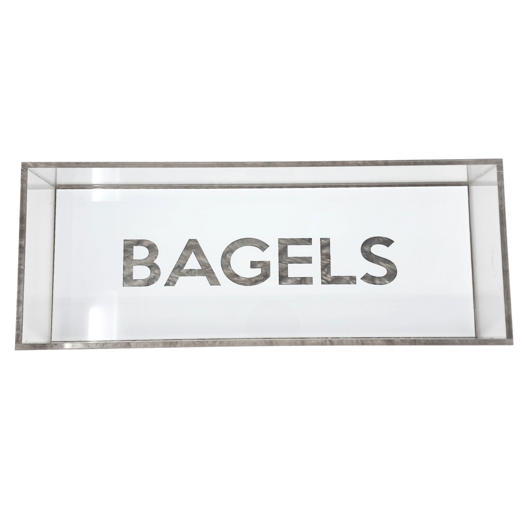 The Eye Candy Bagel Tray is a rectangular glass display case with "BAGELS" in bold letters on the front, a food-safe acrylic tray for bagels, and stylish silver marble acrylic accents.