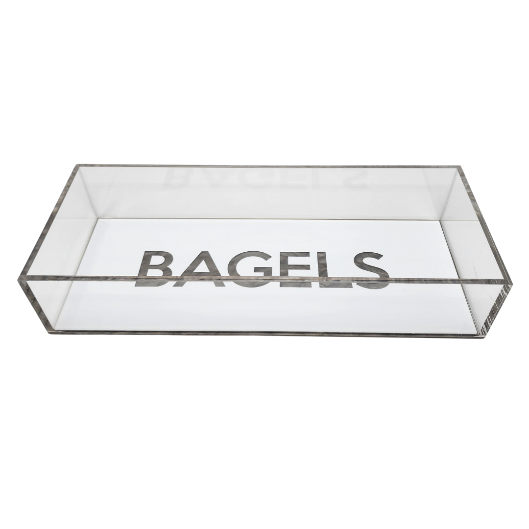The Eye Candy Bagel Tray is a rectangular, clear silver marble acrylic box with "BAGELS" in bold capitals on the inside bottom. Food safe and lidless, it offers a modern touch for serving bagels.
