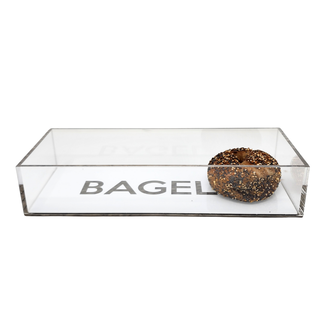 An everything bagel rests in Eye Candy's Bagel Tray—a clear, food-safe silver marble acrylic case with "BAGEL" displayed on the bottom inside.