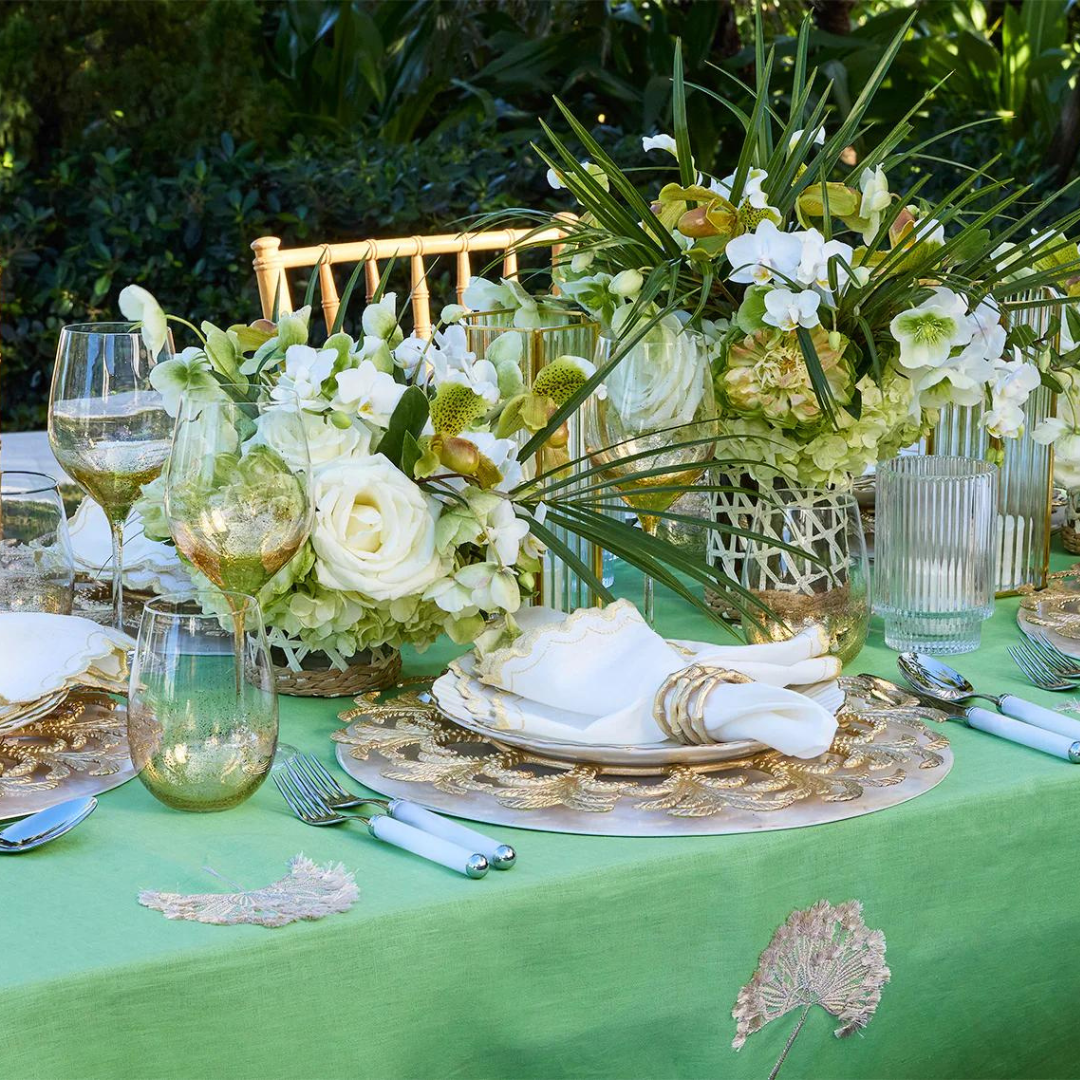 An elegant outdoor table with a green tablecloth, gold-accented glassware, white plates, floral centerpieces, gold chairs, and Kim Seybert Bamboo Napkin Ring Gold Set of 4, all surrounded by lush greenery.