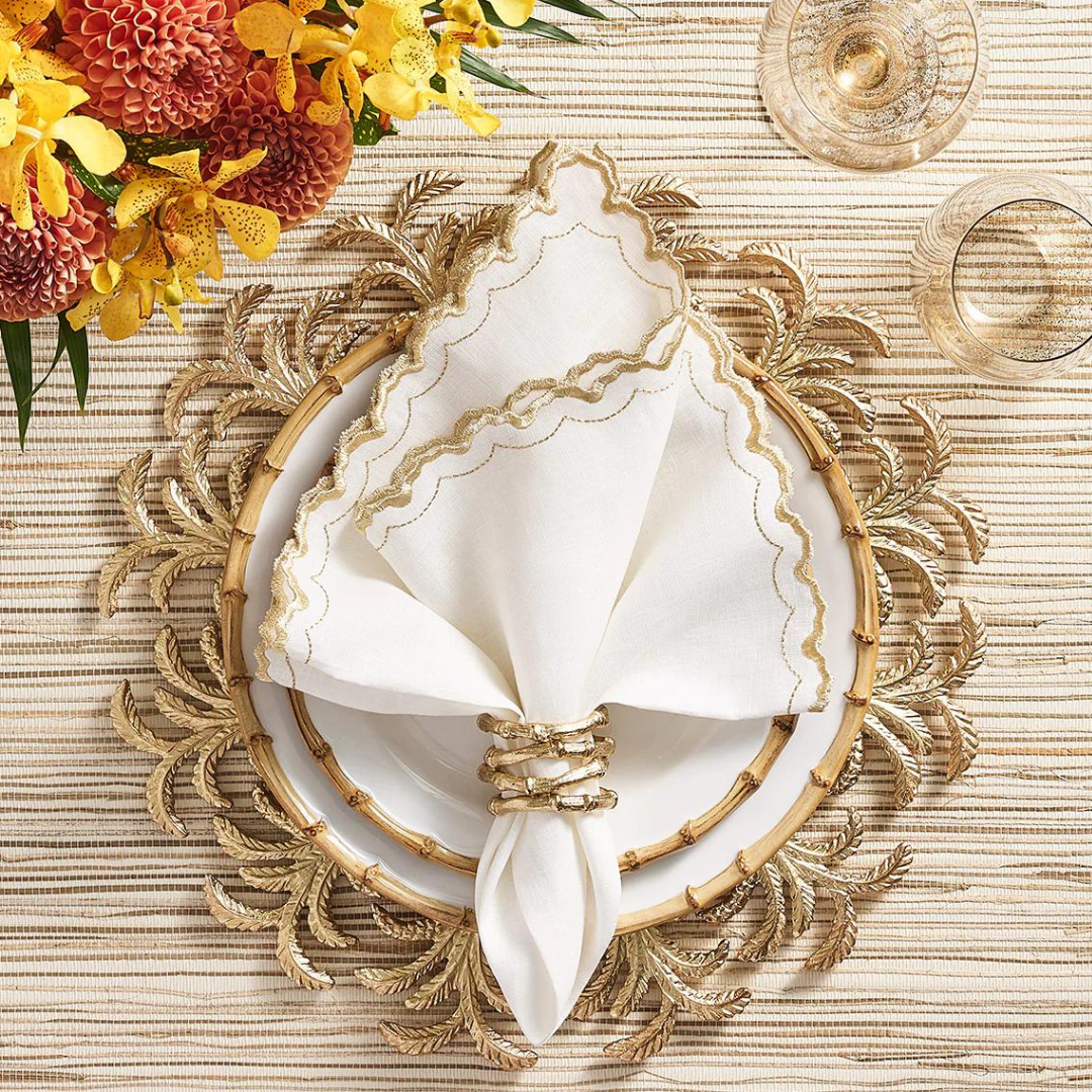 A white plate rests on a gold, leaf-patterned placemat with a white napkin trimmed in gold, secured by a Kim Seybert Bamboo Napkin Ring Gold Set of 4. Yellow and red flowers and glassware complete the table setting.