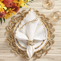 A white plate rests on a gold, leaf-patterned placemat with a white napkin trimmed in gold, secured by a Kim Seybert Bamboo Napkin Ring Gold Set of 4. Yellow and red flowers and glassware complete the table setting.