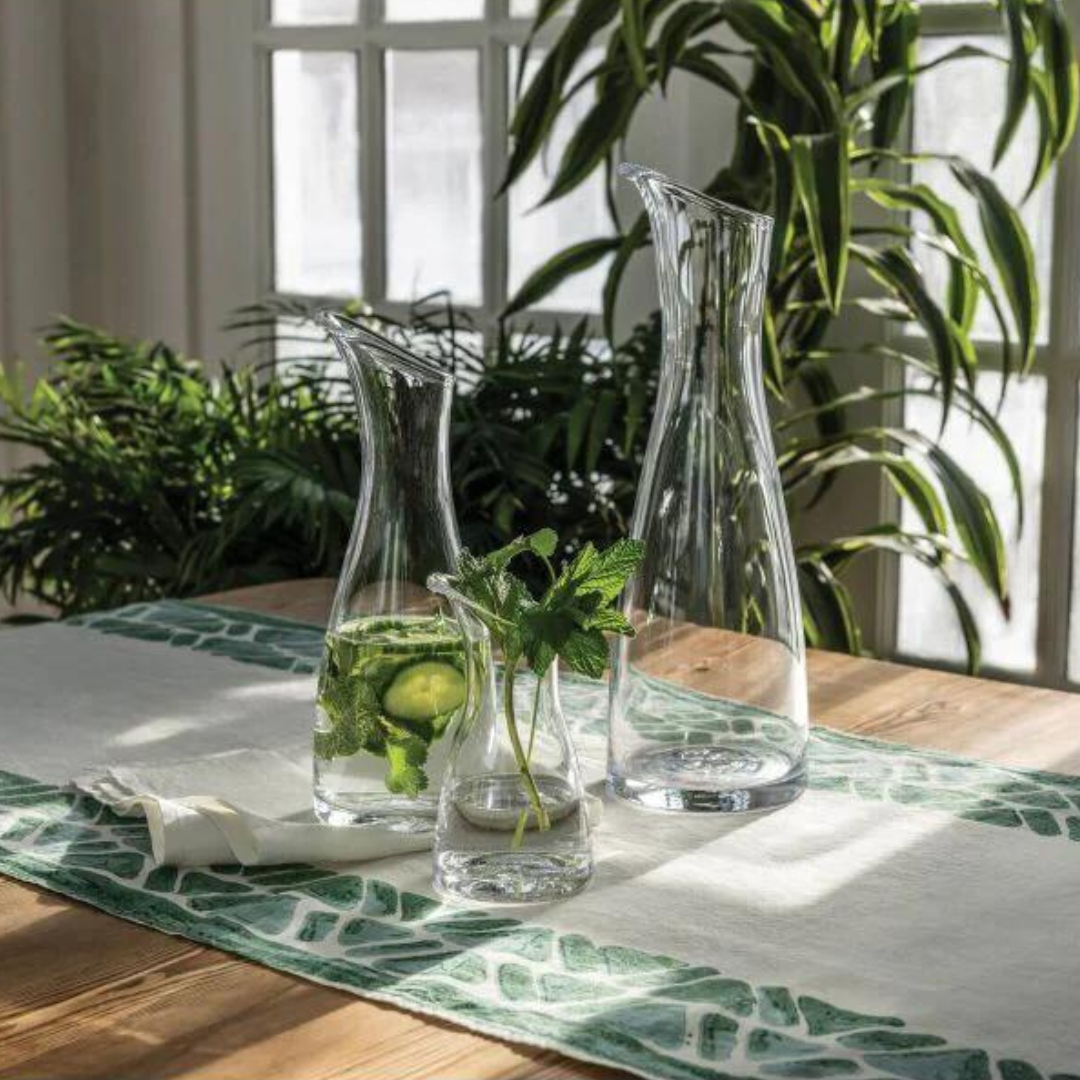 Two Simon Pearce Barre Carafes and a small glass with mint, lemon, and cucumber rest on a wooden table with a green-and-white runner. Sunlight pours through large windows, illuminating lush green plants in the background.