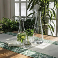 Two Simon Pearce Barre Carafes and a small glass with mint, lemon, and cucumber rest on a wooden table with a green-and-white runner. Sunlight pours through large windows, illuminating lush green plants in the background.