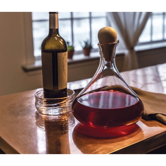 The Simon Pearce Vintner Wine Decanter With Marble Stopper, filled with red wine, stands on a reflective table next to a bottle in a glass dish. Sheer curtains and small potted plants decorate the window behind.