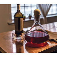 The Simon Pearce Vintner Wine Decanter With Marble Stopper, filled with red wine, stands on a reflective table next to a bottle in a glass dish. Sheer curtains and small potted plants decorate the window behind.