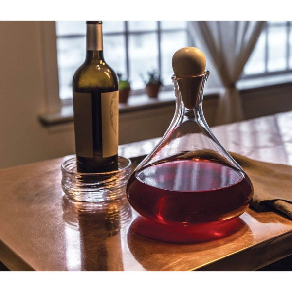 The Simon Pearce Vintner Wine Decanter With Marble Stopper, filled with red wine, stands on a reflective table next to a bottle in a glass dish. Sheer curtains and small potted plants decorate the window behind.