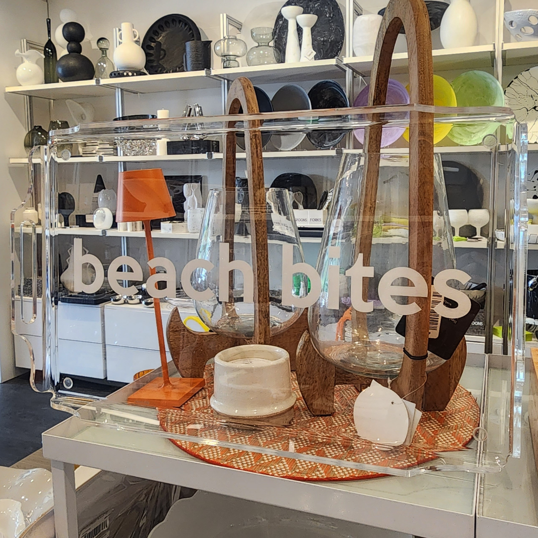 The Eye Candy Deluxe Acrylic Beach Bites Tray displays wooden and glass serveware, a white bowl, an orange tray, and tableware—ideal for outdoor entertaining—in a modern store filled with decorative accents.