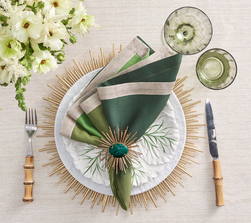 An elegant formal table setting featuring the Kim Seybert Radiate Charger in Gold, a white textured plate, green and linen napkin with jeweled ring, two green glasses, bamboo-handled cutlery, and white flowers in the upper left corner.