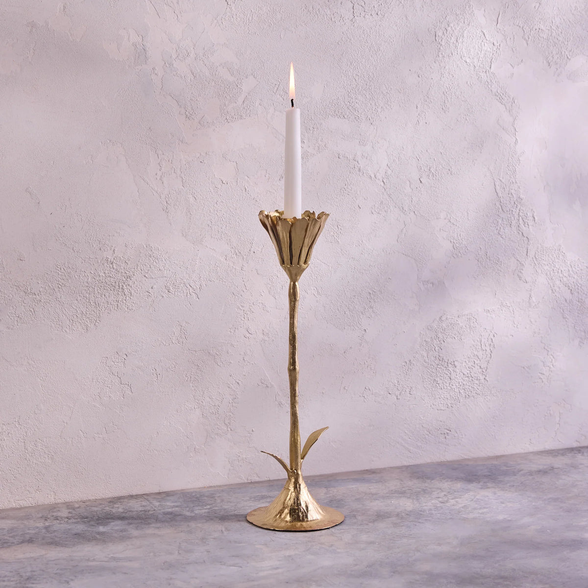 A lit white candle rests in the Be Home Botanic Gold Candleholder, handcrafted in India from brass with a leaf-accented stem. It’s displayed on a gray textured surface against a light textured wall.