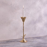 A lit white candle rests in the Be Home Botanic Gold Candleholder, handcrafted in India from brass with a leaf-accented stem. It’s displayed on a gray textured surface against a light textured wall.