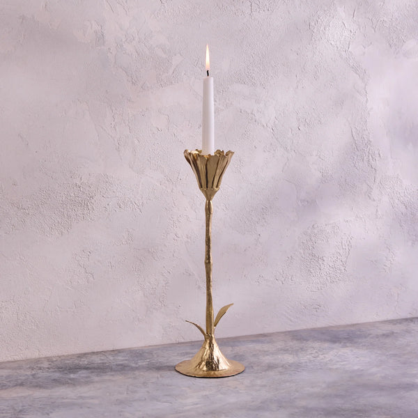 A lit white candle rests in the Be Home Botanic Gold Candleholder, handcrafted in India from brass with a leaf-accented stem. It’s displayed on a gray textured surface against a light textured wall.