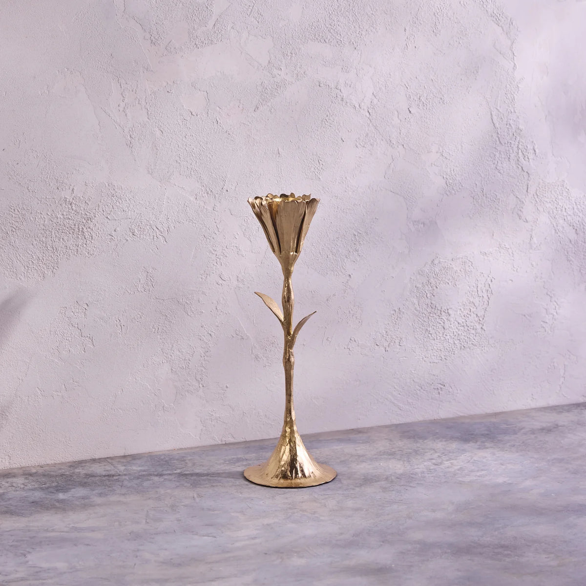 The Botanic Gold Candleholder by Be Home, handmade in India and shaped like a flower stem with leaves, stands on a textured gray surface against a light, textured wall.