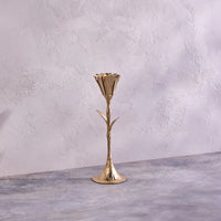 The Botanic Gold Candleholder by Be Home, handmade in India and shaped like a flower stem with leaves, stands on a textured gray surface against a light, textured wall.
