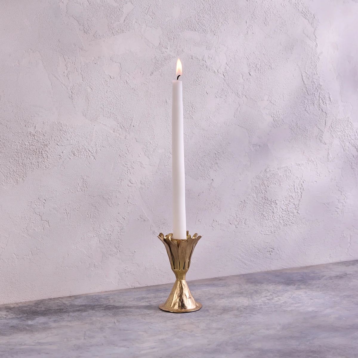 A single white taper candle is lit and placed in the Be Home Botanic Gold Candleholder, handmade in India, on a gray textured surface with a light textured wall in the background.
