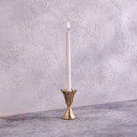 A single white taper candle is lit and placed in the Be Home Botanic Gold Candleholder, handmade in India, on a gray textured surface with a light textured wall in the background.