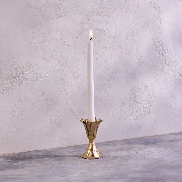 A single white taper candle is lit and placed in the Be Home Botanic Gold Candleholder, handmade in India, on a gray textured surface with a light textured wall in the background.