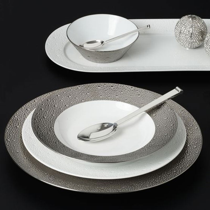 The Bernardaud Ecume Platinum Dinnerware set from Limoges, France features textured silver and white plates, a coordinating bowl, silver spoons, and a decorative oval tray with two silver balls displayed on a modern black surface.