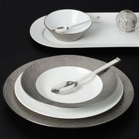 The Bernardaud Ecume Platinum Dinnerware set from Limoges, France features textured silver and white plates, a coordinating bowl, silver spoons, and a decorative oval tray with two silver balls displayed on a modern black surface.