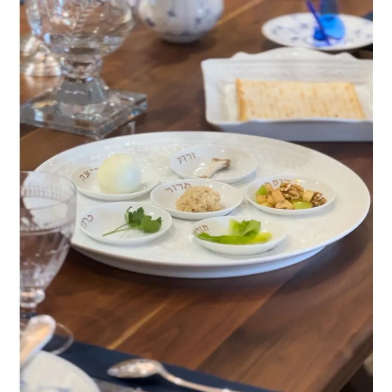 A Louvre Seder Dishes Set of 6 by Bernardaud displays Passover foods—egg, parsley, romaine, charoset, horseradish, and shank bone—on a wooden table set for a meal with matzah in the background.